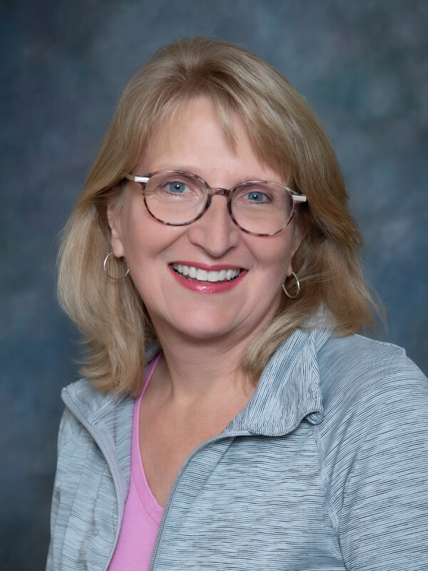 Headshot of Maria McGannon, nurse practitioner at South Lake Pediatrics. Maria has blonde hair, is wearing a grey zip-up top over a pink shirt, and is smiling at the camera.