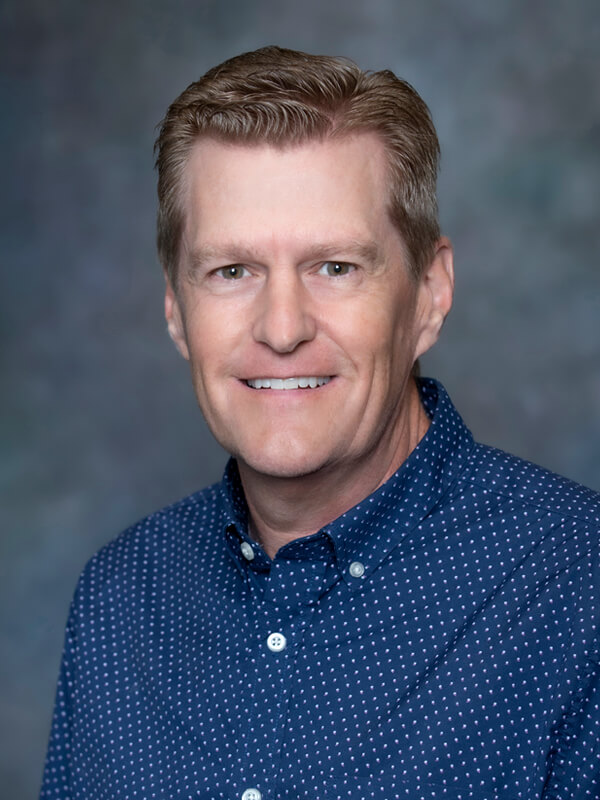 Headshot of John Paulson, pediatrician at South Lake Pediatrics. John has short blonde hair, is wearing a dark blue shirt with small white dots, and is smiling at the camera.